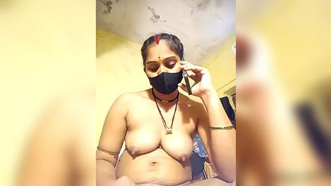 Media: Video of a topless, light-skinned woman with medium-sized breasts, wearing a black face mask, adjusting her hair in a yellow room with a ceiling fan.