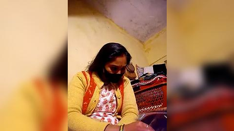 Media: Video of a South Asian woman with medium brown skin, long black hair, and a floral blouse, sitting indoors in a dimly lit room with yellow walls and a wicker chair.