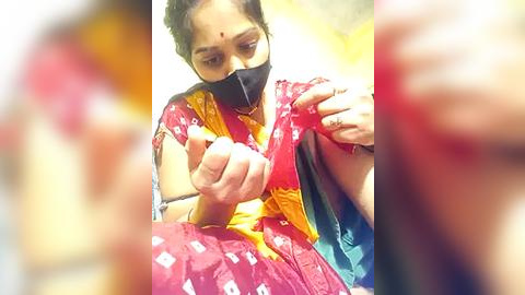 Media: Video of a woman in a colorful sari with a mask, meticulously sewing a garment, with blurred background.