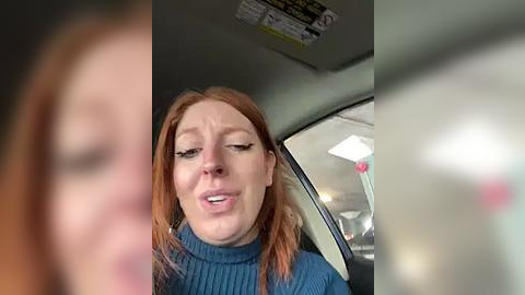 Media: A video of a woman with long red hair and fair skin, wearing a blue turtleneck, appears distressed, her eyes closed, mouth open, inside a car with blurred background.