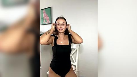Media: Video of a young woman with light skin and long, straight hair, wearing a black one-piece swimsuit, adjusting her hair in a minimalist, white-walled room.