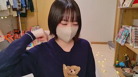 Media: Video of an Asian woman with short black hair and pale skin, wearing a beige face mask, navy sweater with a teddy bear graphic, and a blue surgical glove on her right hand. Background features a cluttered, brightly lit room with toys and clothes.