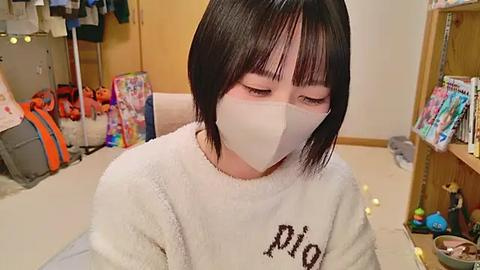 Media: Video of a young Asian woman with short black hair, wearing a white mask, in a cozy, cluttered room with a plush sofa, toys, and a bookshelf.