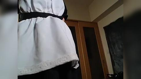 Media: A video of a maid's uniform from the back, featuring a white apron with lace trim, black waistband, and a black skirt. The background shows a wooden door and beige walls.