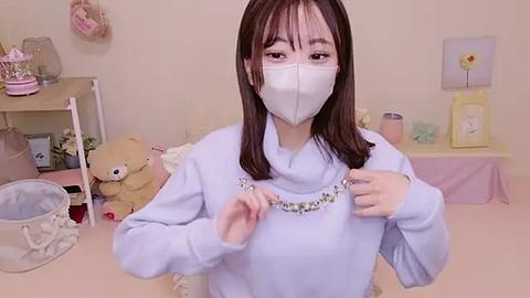 Media: Video of an Asian woman with long brown hair, wearing a white mask, light blue sweater with a floral necklace, and a teddy bear, in a pastel-themed bedroom with floral decor.