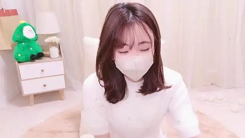 Media: Video of an Asian woman with straight brown hair, wearing a white mask, seated on a beige rug in a minimalist, beige-tiled room with a green plush toy and a lamp on a wooden side table.