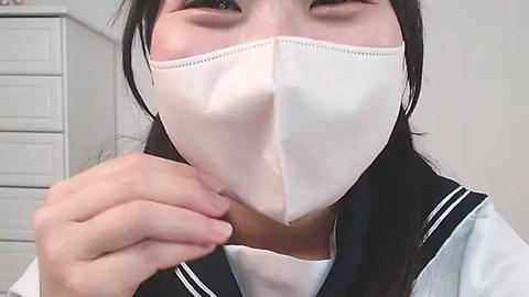 Media: Video of an Asian woman with long black hair, wearing a white surgical mask, black sailor uniform, and white shirt, holding a small object near her mouth, indoors with white drawers in the background.