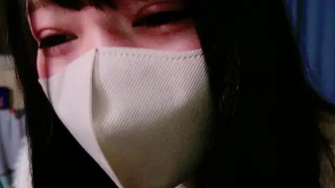 Media: Video of a person with long black hair, wearing a beige medical mask, partially visible face with closed eyes, blurred background.