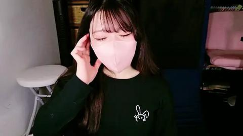 Media: Video of an Asian woman with long black hair, wearing a black top with a bunny logo, a pink face mask, and adjusting her mask with her right hand. Background shows a white stool, a dark shelf with pink items, and a blue cushion.