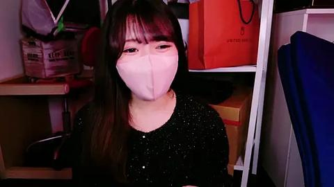 Media: A video of an Asian woman with long black hair and fair skin wearing a white face mask, black glittery top, and seated in a cluttered closet filled with bags and boxes.