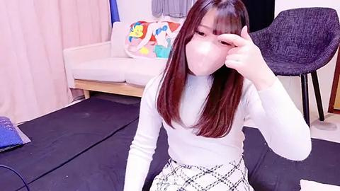 Media: Video of an East Asian woman with long brown hair, wearing a white turtleneck and a black-and-white plaid skirt, sitting on a black mat in a bedroom with a white bed and colorful pillows.