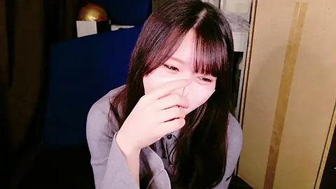 Media: A video of an East Asian woman with long black hair, wearing a gray sweater, covering her nose with her hand, in a dimly lit room with a blue chair and a spherical lamp.