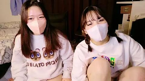 Media: Video of two young Asian women with long brown hair, wearing white sweatshirts with a cartoon cat design and surgical masks, sitting side by side indoors.