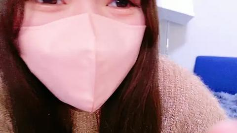 Media: A close-up video of a person wearing a light pink surgical mask, brown hair, and a brown sweater, indoors with white walls and a blue couch in the background.