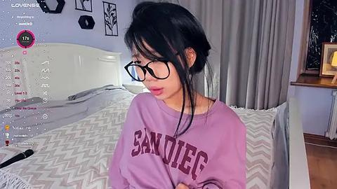 Media: Video of an Asian woman with long black hair, wearing glasses and a purple \"San Diego\" sweatshirt, standing in a bedroom with a white bed and gray curtains.