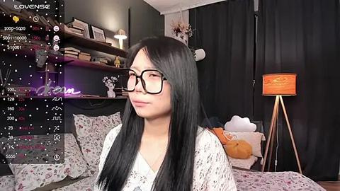 Media: Video of an East Asian woman with long black hair, wearing glasses and a floral-patterned dress, sitting on a bed in a cozy, dimly-lit room with a lamp, shelves, and stuffed animals.