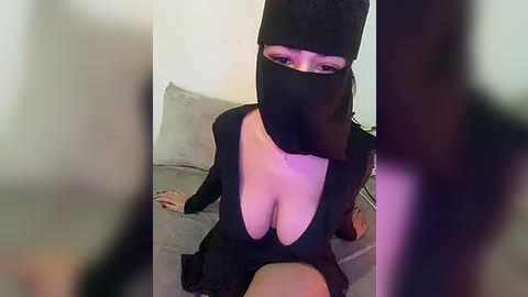 Media: Video of a woman with a black mask, revealing cleavage, sitting on a couch in a dimly lit room.