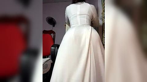 Media: Video of a woman wearing a white, long-sleeved, A-line wedding dress with a high neckline, standing in a room with a red chair and a black tripod.