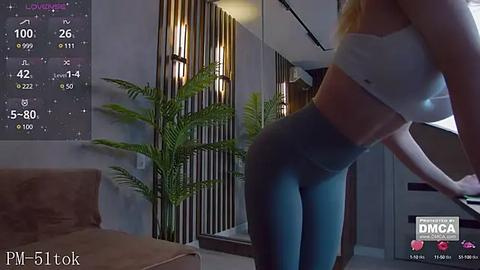 Media: Video of a fit woman with fair skin, long blonde hair, in a white sports bra and blue leggings, bending over, standing in a modern living room with a brown couch, green potted plant, and wooden wall panels.