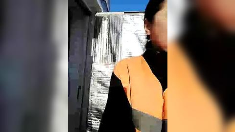 Media: Video of a man in an orange safety vest and black shirt, partially obscured by a reflective, silver barrier, standing in a bright, sunny outdoor setting.