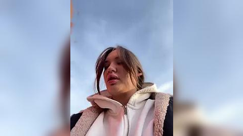 Media: Video of a young woman with light brown hair, wearing a white hoodie and fur-lined jacket, standing against a clear blue sky.