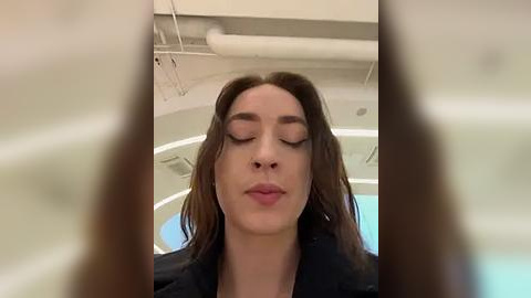 Media: Video of a woman with long brown hair, closed eyes, and lips slightly parted, wearing a black jacket, indoors with a round, white ceiling light and visible air ducts.