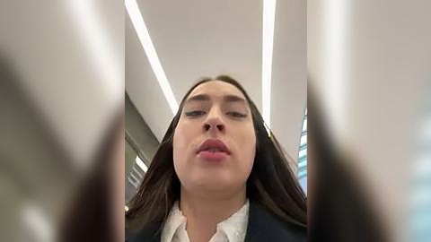 Media: A close-up video of a young woman with long brown hair, wearing a white shirt and dark blazer, looking straight ahead. Background features a modern office interior with white ceiling lights and glass windows.