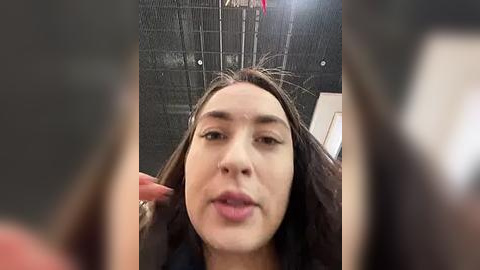 Media: Video of a young woman with long dark hair, light skin, and full lips, taking a selfie indoors with a blurry background.