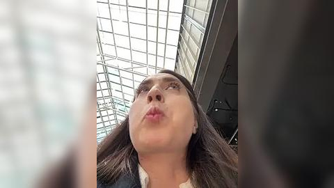 Media: Video of a young woman with long brown hair, wearing a white shirt, looking upward at a glass ceiling with a grid pattern, indoors.