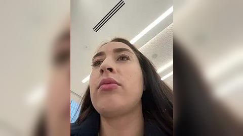 Media: Video of a woman with fair skin and long dark hair, wearing a black coat, taken from a low angle, showing her from the chest up. Background features a modern office setting with white walls and ceiling.