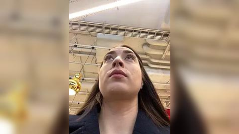 Media: Video of a young woman with fair skin and long brown hair, wearing a black jacket, looking upwards in a ceiling-hung industrial space with yellow light fixtures.