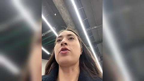 Media: Video of a woman with light brown skin, straight brown hair, and a neutral expression. She wears a dark jacket, standing in an industrial space with exposed pipes and fluorescent lights overhead.