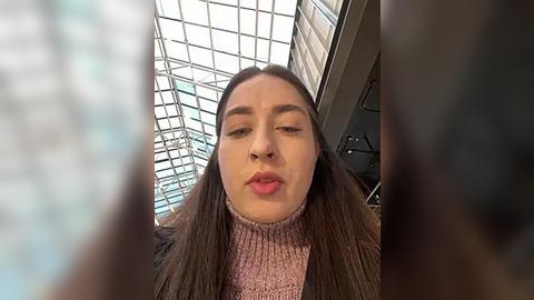 Media: Video of a young woman with long brown hair, wearing a pink turtleneck sweater, standing indoors near a glass-walled atrium with a grid-patterned ceiling.