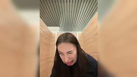 Media: Video of a woman with long dark hair, wearing a black top, peering through a narrow gap between two wooden panels, with a ceiling featuring horizontal metal strips.