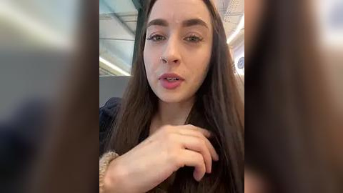 Media: Video of a young woman with fair skin, long brown hair, and a nose ring, wearing a black top, sitting on an airplane. Background shows blurred airplane interior with seats.