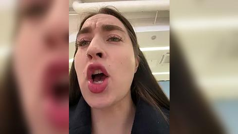 Media: Video of a young woman with long brown hair, fair skin, and pink lips, wearing a black top, appearing shocked or surprised with her mouth open. Background shows a blurred indoor setting with white ceiling and lighting.