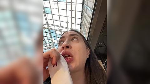 Media: Video of a woman with light skin and long brown hair, wearing a white top, eating a piece of paper, in a modern building with glass walls and a grid ceiling.