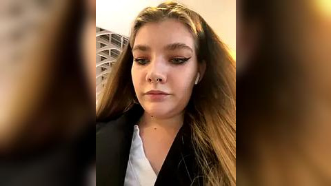 Media: A video of a young woman with light skin, long brown hair, and a nose ring, wearing a black blazer over a white shirt, standing against a blurred modern building background.