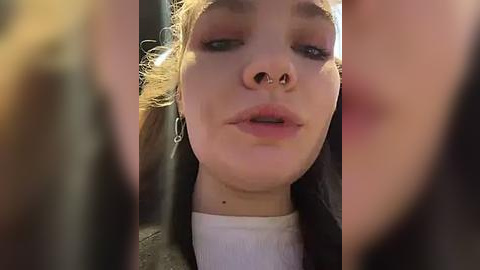 Media: A close-up video of a young woman with fair skin, wearing a black jacket over a white shirt, and a nose ring. Her hair is messy, and her expression is neutral.