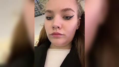 Media: Video of a young woman with fair skin, blue eyes, and light brown hair, wearing a nose ring, black lipstick, and dramatic winged eyeliner. She's in a black jacket over a white shirt, indoors with blurred background.