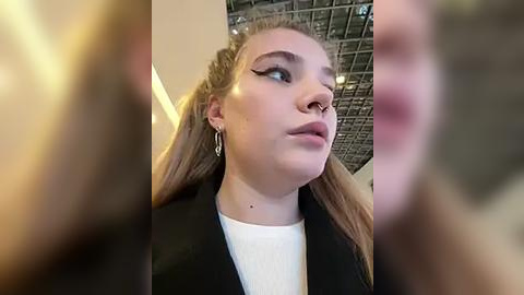 Media: Video of a young woman with light skin and blonde hair, wearing a black blazer over a white shirt, standing indoors with a metallic ceiling.