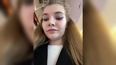 Media: Video of a young woman with light skin, long blonde hair, wearing black clothes, white top, and dark eyeliner. Background is blurred, focusing on her face, nose piercing, and makeup.