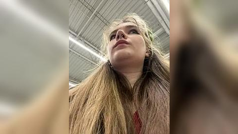 Media: A close-up video of a woman with long, straight, blonde hair and fair skin, wearing a red necklace. She gazes upward, with the ceiling and fluorescent lights visible in the background.