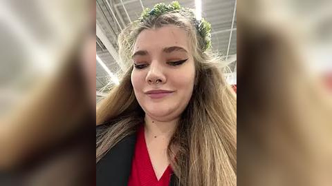 Media: Video of a young woman with long, straight, light brown hair adorned with a green headband. She has a nose ring and is wearing a black jacket over a red top, with her eyes closed and a slight smile.