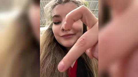 Media: A video of a young woman with long, straight brown hair, wearing a black top, holding her index finger up in front of her face, smiling. Background is blurred.