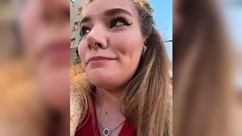 Media: Video of a young woman with fair skin, light brown hair in pigtails, and a nose ring, wearing a red top, smiling outdoors with a blurred background.