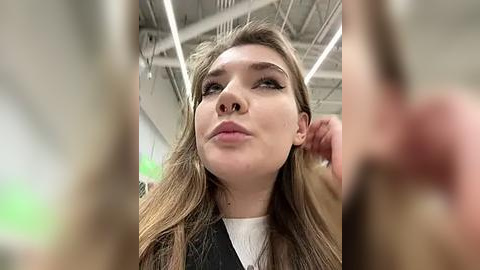 Media: Video of a young Caucasian woman with long, wavy light brown hair, wearing a white shirt and black blazer, looking contemplative in a modern, industrial-style room with exposed ceiling beams and fluorescent lighting.