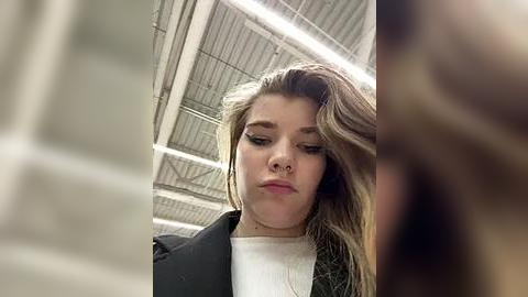 Media: Video of a young woman with long, wavy brown hair, wearing a black blazer over a white shirt, standing in a modern, industrial setting with exposed metal beams and lighting.