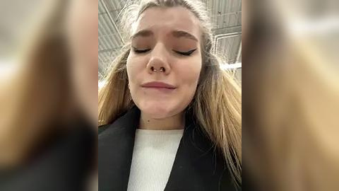 Media: Video of a young woman with long, blonde hair, closed eyes, and a nose ring, wearing a black jacket over a white sweater, inside a modern building with metallic ceiling panels.