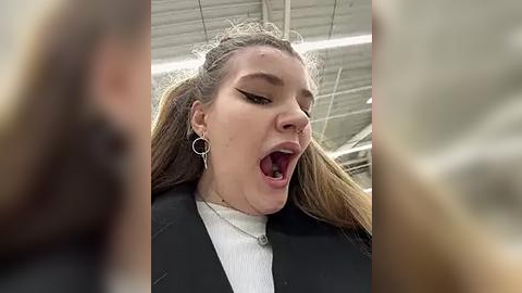 Media: Video of a young woman with long brown hair, wearing a black jacket, white shirt, and silver hoop earrings, mouth open in a dramatic pose. Background shows a blurred industrial setting with exposed pipes.